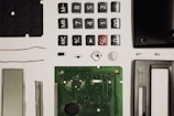 A collection of disassembled electronic components including buttons, a circuit board, a screen, wires, screws, and other small parts, neatly arranged on a white surface.