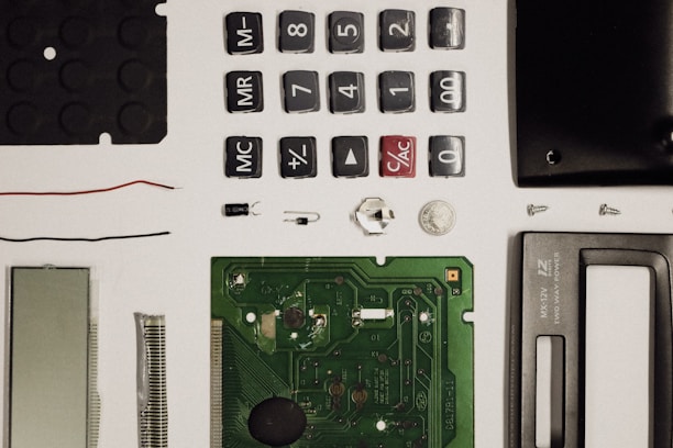 A collection of disassembled electronic components including buttons, a circuit board, a screen, wires, screws, and other small parts, neatly arranged on a white surface.