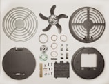 Various disassembled components of a fan displayed neatly on a flat surface. The parts include fan blades, protective grilles, electrical coils, small screws, electronic circuit board, and other mechanical pieces, all arranged in symmetrical fashion.