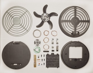 Various disassembled components of a fan displayed neatly on a flat surface. The parts include fan blades, protective grilles, electrical coils, small screws, electronic circuit board, and other mechanical pieces, all arranged in symmetrical fashion.