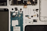 This image features a disassembled smartphone with various components laid out on a flat surface. Visible are the screen, battery, circuit board, camera module, and several smaller components such as screws and connectors. The arrangement is organized and methodical, displaying each part individually.