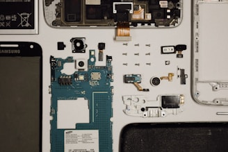 This image features a disassembled smartphone with various components laid out on a flat surface. Visible are the screen, battery, circuit board, camera module, and several smaller components such as screws and connectors. The arrangement is organized and methodical, displaying each part individually.