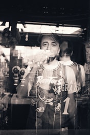 A person stands behind a reflective glass, wearing a basketball jersey with the name McGrady and number 1. The reflection shows both the front and back of the jersey, blending the figure with the surrounding environment. The setting appears to be indoors with a window or light source contributing to the reflections.