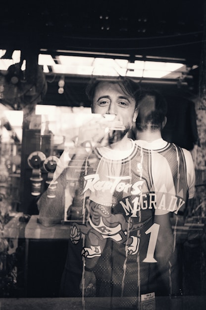 A person stands behind a reflective glass, wearing a basketball jersey with the name McGrady and number 1. The reflection shows both the front and back of the jersey, blending the figure with the surrounding environment. The setting appears to be indoors with a window or light source contributing to the reflections.