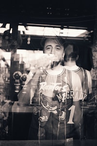 A person stands behind a reflective glass, wearing a basketball jersey with the name McGrady and number 1. The reflection shows both the front and back of the jersey, blending the figure with the surrounding environment. The setting appears to be indoors with a window or light source contributing to the reflections.
