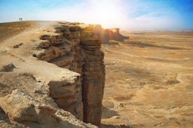 A vast desert landscape with a dramatic cliff edge, illuminated by bright sunlight. Two small figures stand atop the cliff, emphasizing the scale and grandeur of the surroundings. The terrain is composed of layers of rock formations and sprawling sand dunes extending into the horizon.