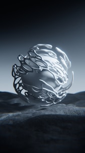 A futuristic, abstract metallic sculpture with flowing, organic shapes stands on a textured surface. The lighting casts intricate shadows, enhancing the three-dimensional appearance.