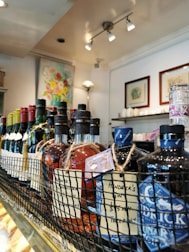 A close-up view of various bottles of liquor and wine displayed on a shelf. The bottles are arranged in a row within a wire basket, with visible labels and tags attached to them. Behind the drinks, a brightly lit interior with artworks and decor can be seen.
