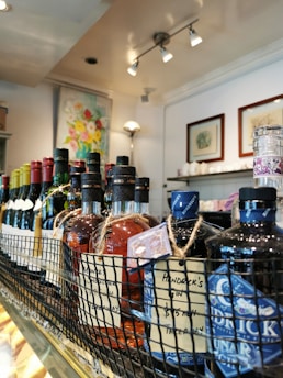 A close-up view of various bottles of liquor and wine displayed on a shelf. The bottles are arranged in a row within a wire basket, with visible labels and tags attached to them. Behind the drinks, a brightly lit interior with artworks and decor can be seen.