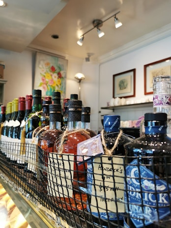 A close-up view of various bottles of liquor and wine displayed on a shelf. The bottles are arranged in a row within a wire basket, with visible labels and tags attached to them. Behind the drinks, a brightly lit interior with artworks and decor can be seen.