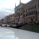 A gondola with passengers is gliding through a canal lined with historic Venetian buildings. The gondolier, wearing a striped shirt, is navigating the boat. The water reflects the sky, which is overcast with patches of blue and clouds.