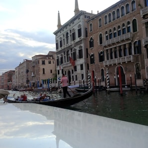A gondola with passengers is gliding through a canal lined with historic Venetian buildings. The gondolier, wearing a striped shirt, is navigating the boat. The water reflects the sky, which is overcast with patches of blue and clouds.