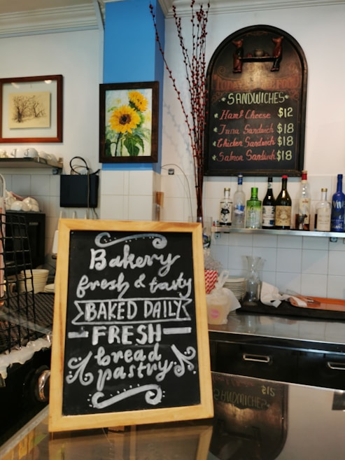 A cozy cafe interior features a blackboard sign with handwritten text advertising fresh bakery items, including bread and pastries. Above the sign, a menu board lists various sandwiches with corresponding prices. The walls are adorned with framed art, including a bright sunflower painting. Several bottles of wine and spirits are lined up on a shelf, and various kitchen items are visible, suggesting a welcoming and artisanal atmosphere.