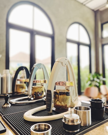 A modern coffee bar setup with espresso machines, tampers, and various coffee accessories arranged on a mat. The background features large arched windows letting in natural light, adding to the airy atmosphere.