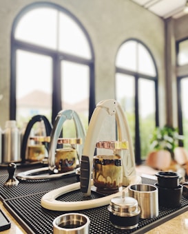 A modern coffee bar setup with espresso machines, tampers, and various coffee accessories arranged on a mat. The background features large arched windows letting in natural light, adding to the airy atmosphere.