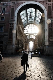 A grand architectural structure with a high, arched glass ceiling allows sunlight to stream through, casting shadows on a cobblestone pathway. People walk along the path, with one prominent individual in a suit carrying a briefcase, heading toward the sunlight.