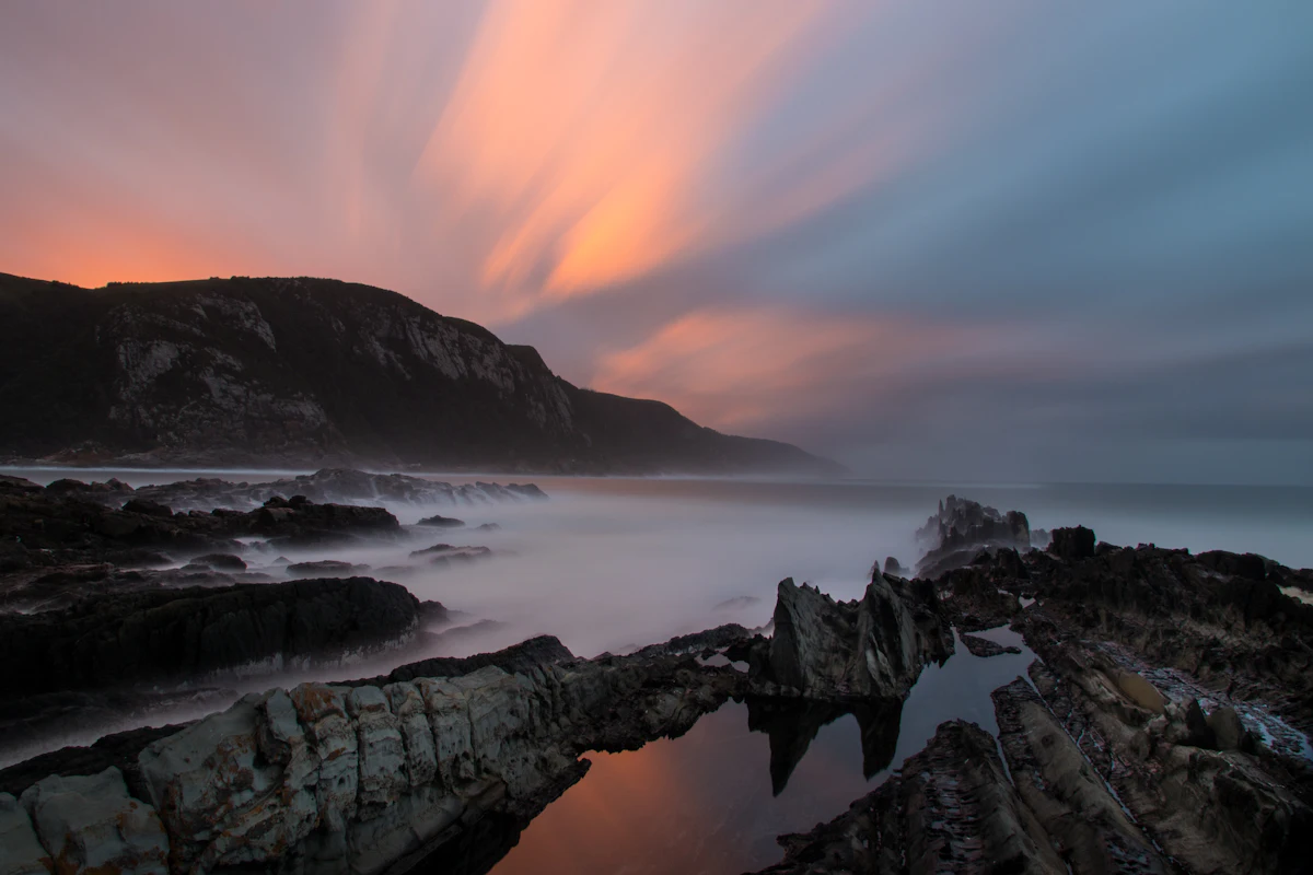 Waves washing over dark rocks at Storms River Mouth at sunrise, with a headland behind