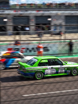 A vibrant green racing car with visible decals and advertisements is in motion on a race track. The backdrop includes another car and blurred spectators behind a chain-link fence.