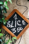 A small chalkboard with a wooden frame displays the text '1 SLICE $3.50' written in white chalk. The board is surrounded by green leaves and set against a textured beige wall, with a black line or cord in the background.
