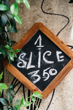 A small chalkboard with a wooden frame displays the text '1 SLICE $3.50' written in white chalk. The board is surrounded by green leaves and set against a textured beige wall, with a black line or cord in the background.