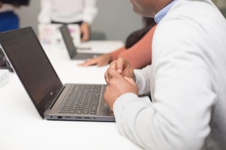 A group of people sitting at a white table using laptops. The focus is on a person in the foreground wearing a light gray sweater, whose hands are visible near the laptop. Another person in a pink top and another individual in the background are partially visible. The setting suggests a collaborative or work-related environment.
