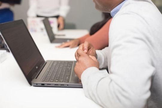 A group of people sitting at a white table using laptops. The focus is on a person in the foreground wearing a light gray sweater, whose hands are visible near the laptop. Another person in a pink top and another individual in the background are partially visible. The setting suggests a collaborative or work-related environment.
