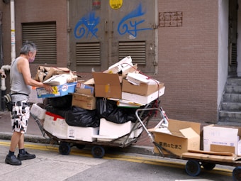 A person wearing a face mask and casual clothing is seen pushing carts filled with cardboard boxes and black trash bags. The scene is set in an urban alleyway with a concrete wall, metal doors, and graffiti. The person appears to be organizing or adjusting the boxes on the carts.