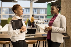 Duas mulheres conversando no ambiente de trabalho, uma está segurando um computador e a outra uma xícara de café