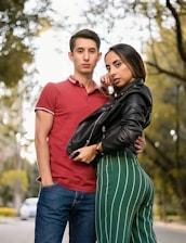 A young couple poses outdoors on a tree-lined path. The man is wearing a red polo shirt and blue jeans, while the woman is in a black leather jacket and green striped pants. They appear confident and stylish, standing closely together with relaxed expressions.