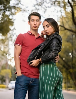 A young couple poses outdoors on a tree-lined path. The man is wearing a red polo shirt and blue jeans, while the woman is in a black leather jacket and green striped pants. They appear confident and stylish, standing closely together with relaxed expressions.