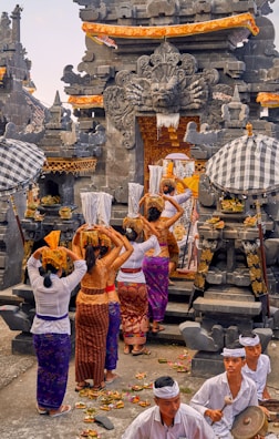 A group of people dressed in traditional attire are participating in a cultural or religious ceremony at a temple. The temple features intricate stone carvings and ornate decorations. Participants are carrying offerings or items on their heads, and musicians are present, playing instruments. The scene is lively and full of cultural significance.