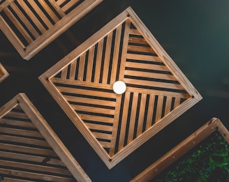 Wooden geometric structures with slatted patterns form symmetrical shapes against a dark background. A central light bulb illuminates the arrangement, creating a warm and structured aesthetic.