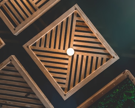 Wooden geometric structures with slatted patterns form symmetrical shapes against a dark background. A central light bulb illuminates the arrangement, creating a warm and structured aesthetic.