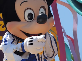 A large, cartoonish character with a smiling face wears a white outfit with blue and gold trim, surrounded by multicolored ribbons against a bright sky.