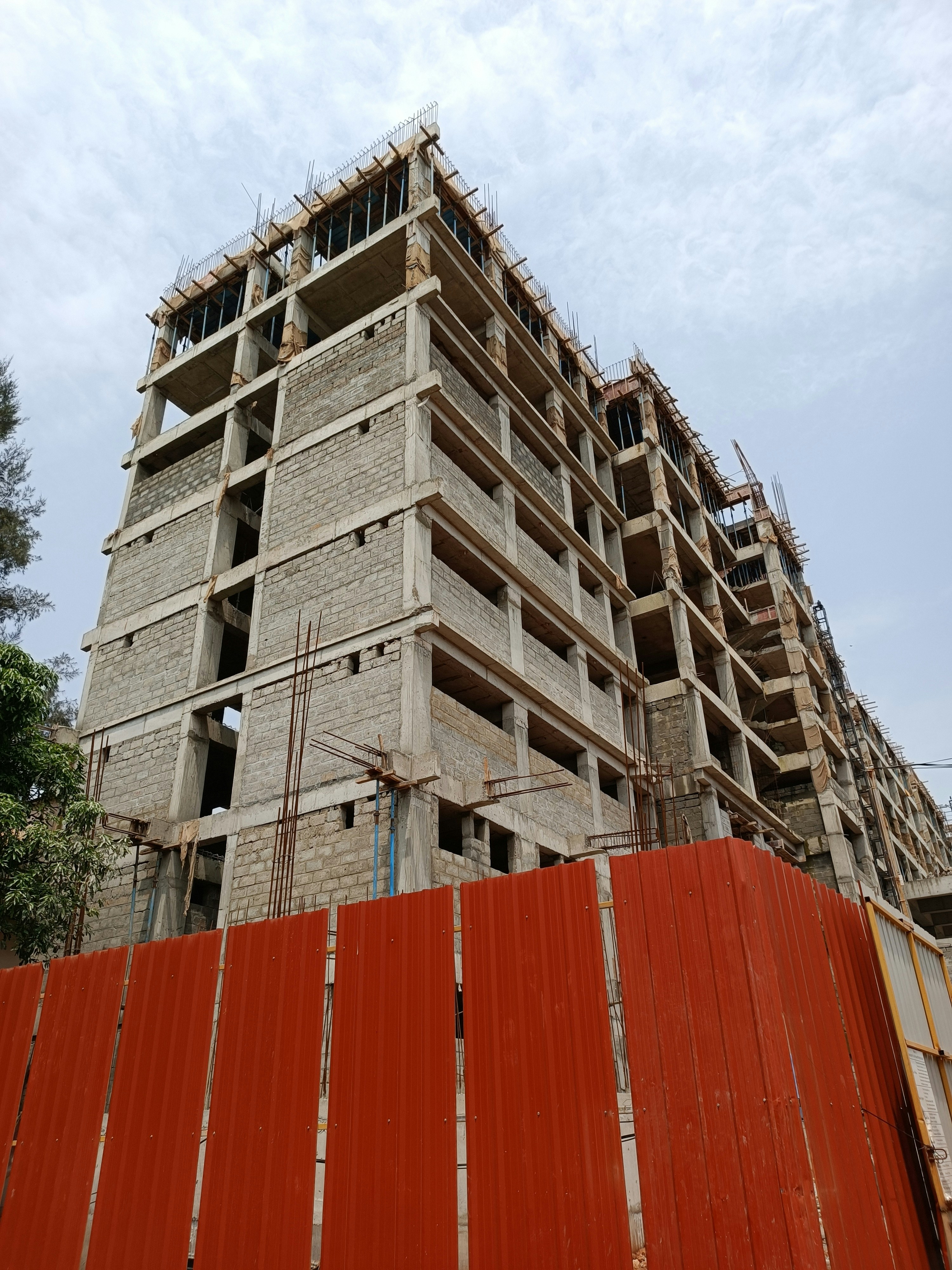 A concrete building under construction with multiple floors is visible. Rebar protrudes from the top, indicating ongoing construction work. A red metal fence surrounds the site, indicating a restricted area. The sky is overcast, adding a neutral tone to the scene.