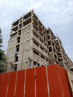 A concrete building under construction with multiple floors is visible. Rebar protrudes from the top, indicating ongoing construction work. A red metal fence surrounds the site, indicating a restricted area. The sky is overcast, adding a neutral tone to the scene.