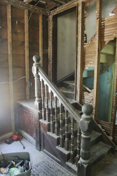 A partially demolished staircase area with exposed wooden framework and old, dusty wooden banisters. The walls are stripped, revealing wooden beams and damaged plaster. A variety of tools and construction materials are scattered on the floor, including a container filled with tools. The scene appears to be in an unfinished renovation state.