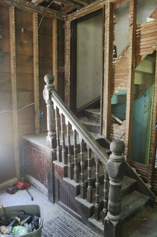 A partially demolished staircase area with exposed wooden framework and old, dusty wooden banisters. The walls are stripped, revealing wooden beams and damaged plaster. A variety of tools and construction materials are scattered on the floor, including a container filled with tools. The scene appears to be in an unfinished renovation state.