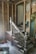 A partially demolished staircase area with exposed wooden framework and old, dusty wooden banisters. The walls are stripped, revealing wooden beams and damaged plaster. A variety of tools and construction materials are scattered on the floor, including a container filled with tools. The scene appears to be in an unfinished renovation state.