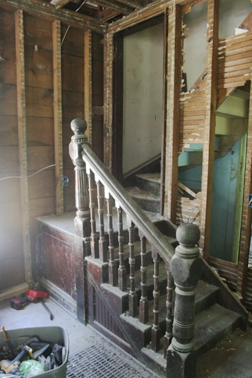 A partially demolished staircase area with exposed wooden framework and old, dusty wooden banisters. The walls are stripped, revealing wooden beams and damaged plaster. A variety of tools and construction materials are scattered on the floor, including a container filled with tools. The scene appears to be in an unfinished renovation state.