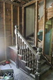 A partially demolished staircase area with exposed wooden framework and old, dusty wooden banisters. The walls are stripped, revealing wooden beams and damaged plaster. A variety of tools and construction materials are scattered on the floor, including a container filled with tools. The scene appears to be in an unfinished renovation state.