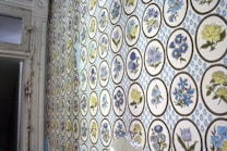 A wall is covered with vintage wallpaper featuring circular motifs containing various floral designs. The flowers are primarily in shades of blue and yellow, set against a cream-colored background. The overall pattern repeats consistently across the wall, giving it a retro aesthetic.