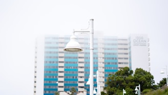 A white street lamp stands in the foreground with a tall multi-story building behind it. The building has blue and white vertical strips and is partially obscured by fog or mist. The words '1900 Ocean Luxury Living' are visible on the building. There are trees and additional street lamps in the background.
