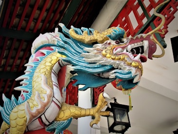 A colorful and ornate dragon sculpture is depicted, featuring intricate details and a vibrant palette. The sculpture has a prominent dragon head with open mouth displaying sharp teeth and whiskers. Its scales are painted in hues of turquoise, gold, and white, with additional decorative elements. The background shows parts of architectural elements with Asian design influences, such as red and green wooden beams and a traditional lantern.