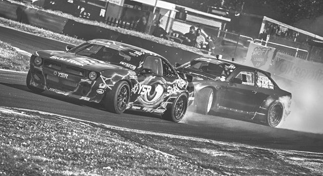 Two cars are drifting on a racetrack, creating a cloud of smoke behind them. The vehicles display various sponsor stickers and are captured in a high-speed action. The image has a dynamic and intense atmosphere, reflecting the competitive nature of drifting.