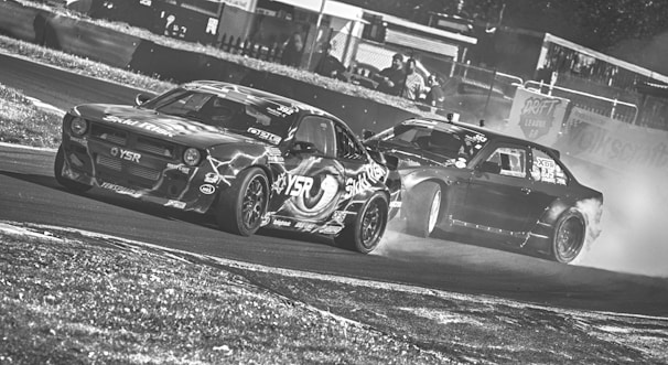 Two cars are drifting on a racetrack, creating a cloud of smoke behind them. The vehicles display various sponsor stickers and are captured in a high-speed action. The image has a dynamic and intense atmosphere, reflecting the competitive nature of drifting.
