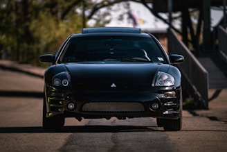 A sleek, black sports car is parked on a quiet, shaded road. The car, likely a Mitsubishi, displays a shiny, well-maintained surface with a front-facing perspective revealing its headlights and grille. Surrounding the car, the blurry background features green foliage and a structure, suggesting an outdoor or suburban setting.