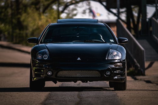A sleek, black sports car is parked on a quiet, shaded road. The car, likely a Mitsubishi, displays a shiny, well-maintained surface with a front-facing perspective revealing its headlights and grille. Surrounding the car, the blurry background features green foliage and a structure, suggesting an outdoor or suburban setting.