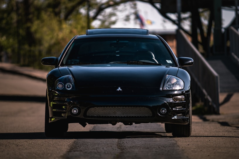 A sleek, black sports car is parked on a quiet, shaded road. The car, likely a Mitsubishi, displays a shiny, well-maintained surface with a front-facing perspective revealing its headlights and grille. Surrounding the car, the blurry background features green foliage and a structure, suggesting an outdoor or suburban setting.