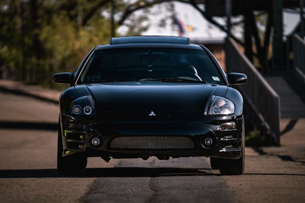 A sleek, black sports car is parked on a quiet, shaded road. The car, likely a Mitsubishi, displays a shiny, well-maintained surface with a front-facing perspective revealing its headlights and grille. Surrounding the car, the blurry background features green foliage and a structure, suggesting an outdoor or suburban setting.